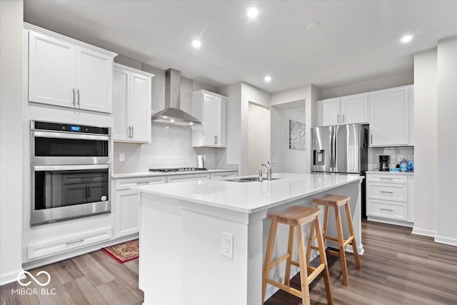 kitchen with wall chimney exhaust hood, stainless steel appliances, a kitchen island with sink, sink, and white cabinets