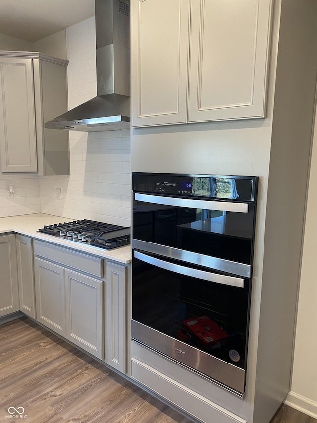 kitchen featuring wall chimney range hood, hardwood / wood-style flooring, tasteful backsplash, stainless steel double oven, and gas cooktop