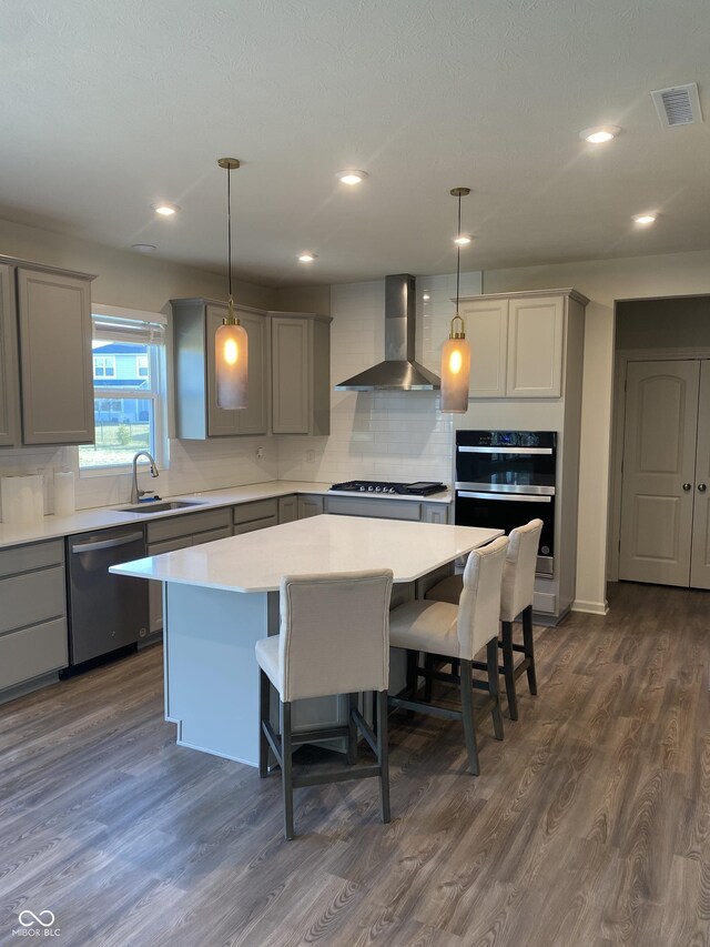 kitchen with dark wood-type flooring, wall chimney range hood, appliances with stainless steel finishes, decorative light fixtures, and a kitchen island