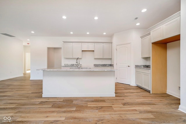 kitchen featuring light wood-type flooring, light stone counters, a kitchen island with sink, sink, and white cabinetry