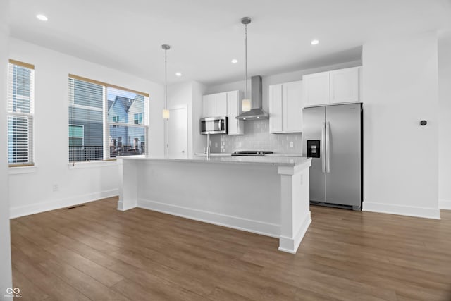 kitchen featuring a kitchen island with sink, wall chimney range hood, decorative backsplash, white cabinetry, and stainless steel appliances