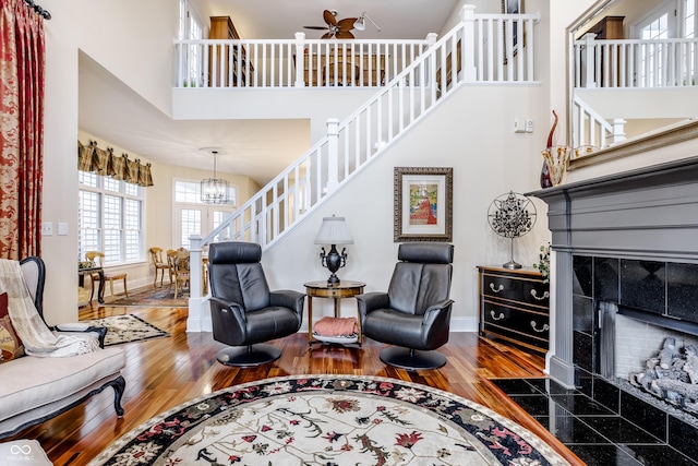 living room with ceiling fan with notable chandelier, a high ceiling, a tiled fireplace, and hardwood / wood-style floors