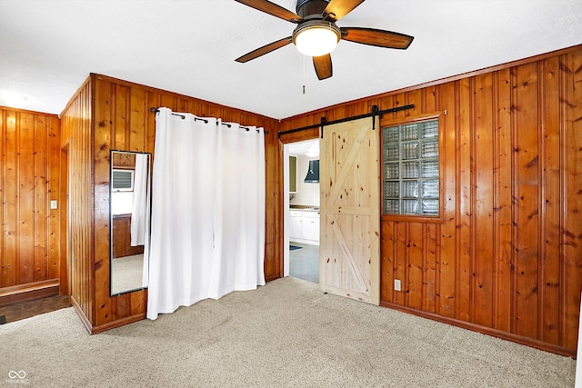 unfurnished bedroom with wood walls, ceiling fan, a barn door, and light colored carpet