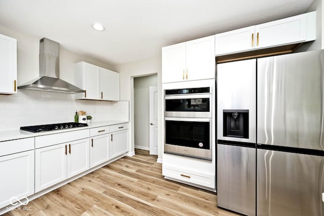 kitchen featuring white cabinets, wall chimney range hood, light hardwood / wood-style flooring, appliances with stainless steel finishes, and tasteful backsplash