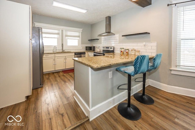 kitchen featuring wall chimney range hood, light stone countertops, appliances with stainless steel finishes, kitchen peninsula, and a breakfast bar area