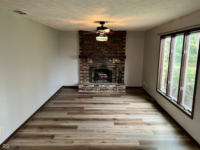 unfurnished living room with a textured ceiling, a ceiling fan, a brick fireplace, visible vents, and wood finished floors