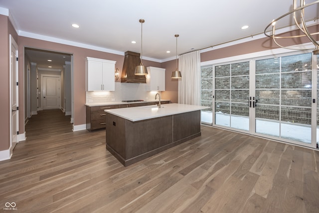 kitchen with custom exhaust hood, hanging light fixtures, hardwood / wood-style floors, a kitchen island with sink, and white cabinetry