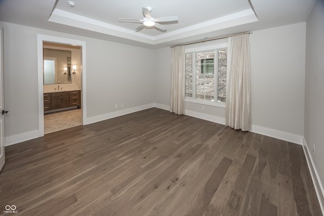 unfurnished bedroom featuring ceiling fan, dark wood-type flooring, a tray ceiling, sink, and ensuite bathroom