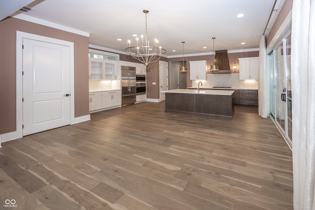 kitchen with stainless steel appliances, white cabinetry, an island with sink, hanging light fixtures, and exhaust hood