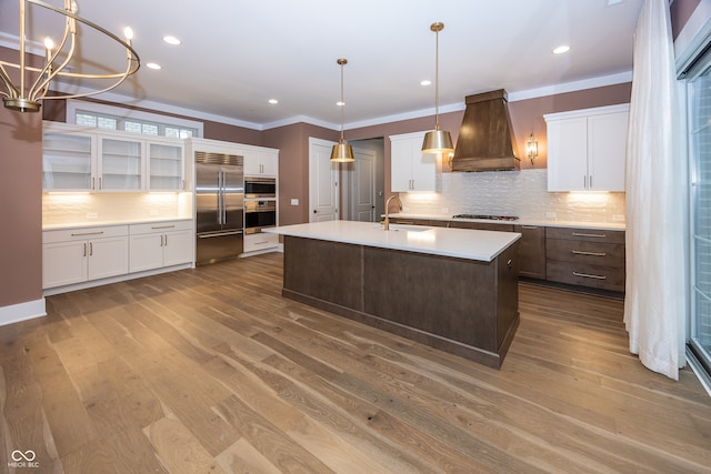 kitchen with a kitchen island with sink, built in refrigerator, custom range hood, sink, and white cabinetry