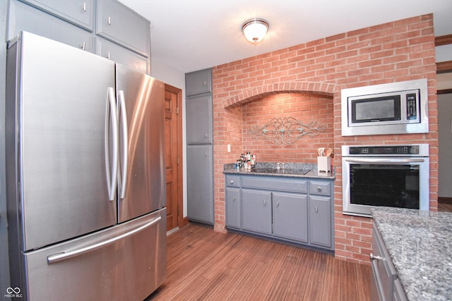 kitchen with appliances with stainless steel finishes, dark stone counters, gray cabinetry, brick wall, and light hardwood / wood-style floors