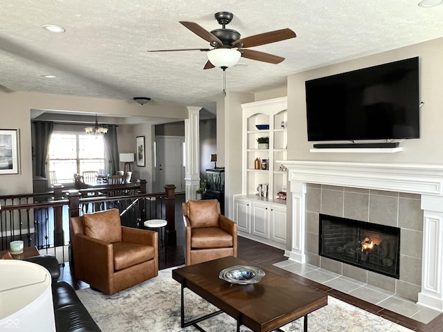 living room featuring ceiling fan with notable chandelier, built in shelves, a textured ceiling, wood-type flooring, and a tiled fireplace