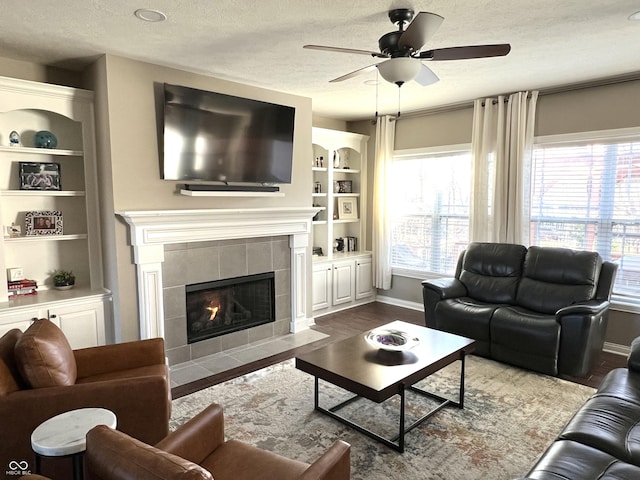 living room with built in shelves, a textured ceiling, ceiling fan, a tile fireplace, and hardwood / wood-style floors