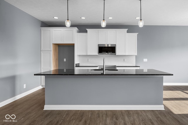 kitchen with dark wood-type flooring, white cabinets, decorative backsplash, stainless steel microwave, and a center island with sink