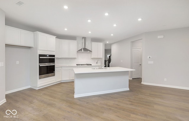 kitchen with light wood-type flooring, double oven, a kitchen island with sink, wall chimney range hood, and white cabinets