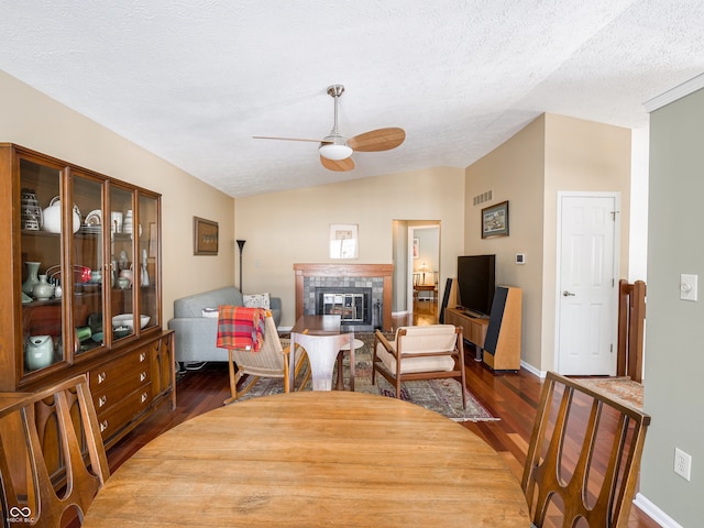 dining room featuring wood finished floors, a ceiling fan, vaulted ceiling, a textured ceiling, and a tiled fireplace