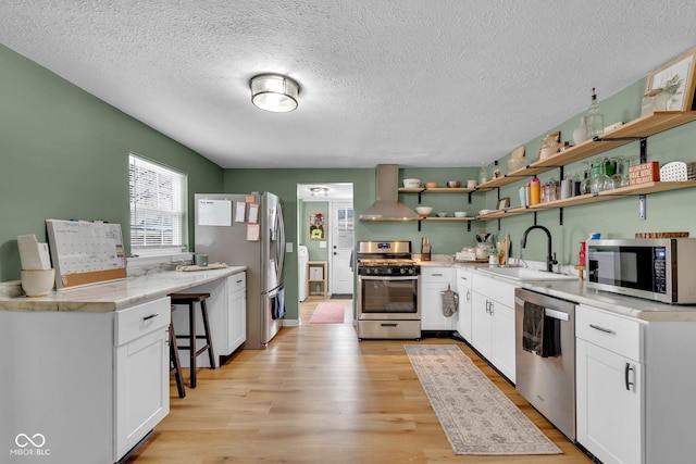 kitchen featuring open shelves, stainless steel appliances, a sink, light wood-type flooring, and wall chimney exhaust hood