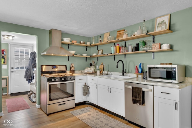 kitchen with light wood finished floors, white cabinets, wall chimney exhaust hood, appliances with stainless steel finishes, and a sink