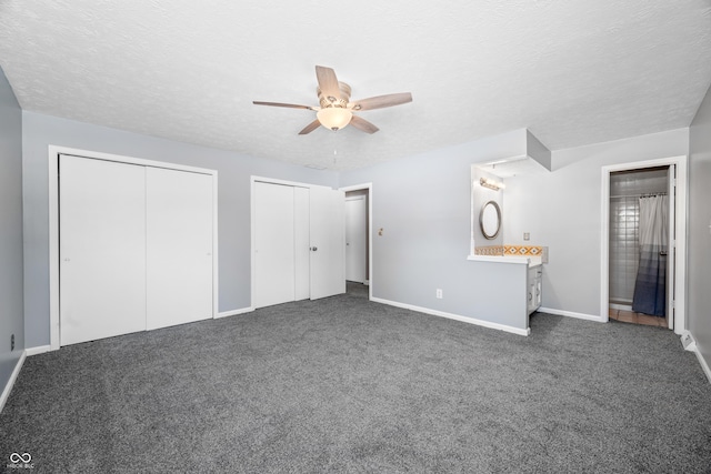 unfurnished bedroom featuring ceiling fan, ensuite bathroom, a textured ceiling, dark carpet, and two closets