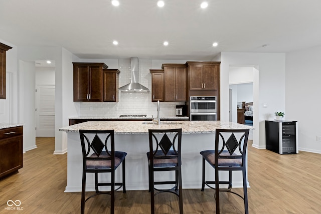 kitchen featuring wine cooler, a kitchen island with sink, stainless steel appliances, wall chimney range hood, and light wood-type flooring