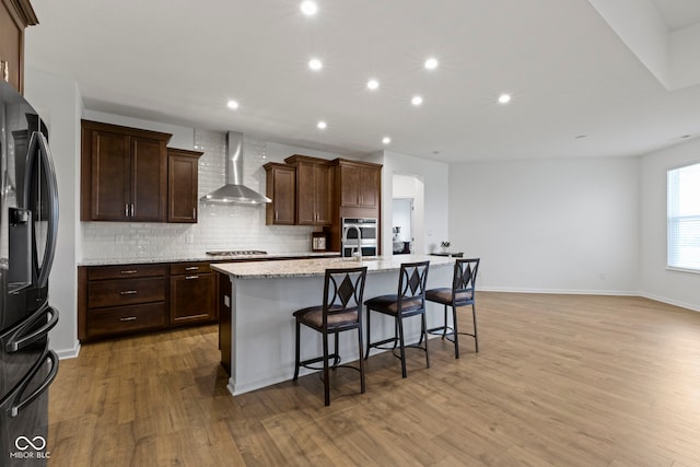 kitchen with light wood-style flooring, black fridge with ice dispenser, wall chimney range hood, a center island with sink, and stainless steel gas stovetop