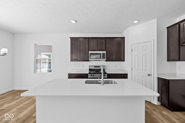 kitchen with a kitchen island with sink, light wood-style flooring, dark brown cabinetry, a sink, and appliances with stainless steel finishes