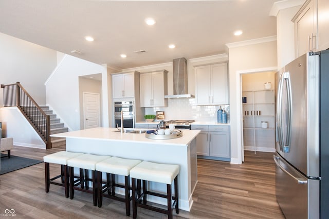 kitchen with a breakfast bar, wall chimney exhaust hood, gray cabinets, an island with sink, and stainless steel appliances