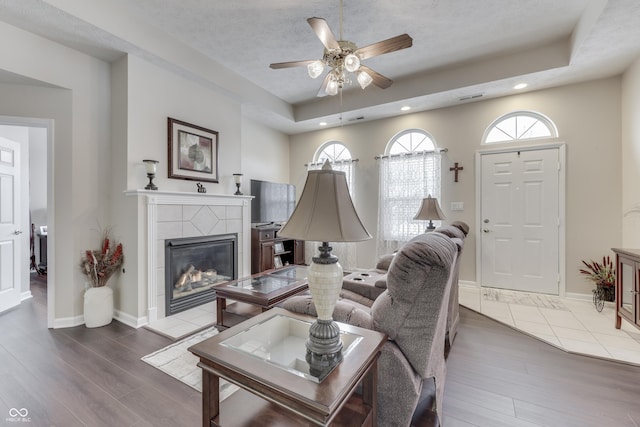 living room with ceiling fan, wood-type flooring, a tile fireplace, and a tray ceiling