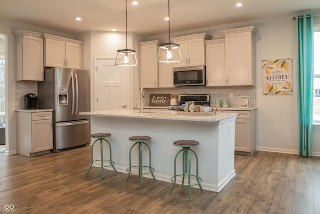 kitchen featuring dark wood-type flooring, hanging light fixtures, tasteful backsplash, an island with sink, and appliances with stainless steel finishes