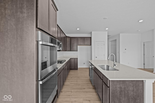 kitchen featuring an island with sink, a sink, dark brown cabinetry, light wood-style floors, and appliances with stainless steel finishes