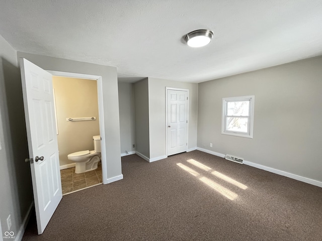 unfurnished bedroom with visible vents, a textured ceiling, connected bathroom, dark colored carpet, and baseboards