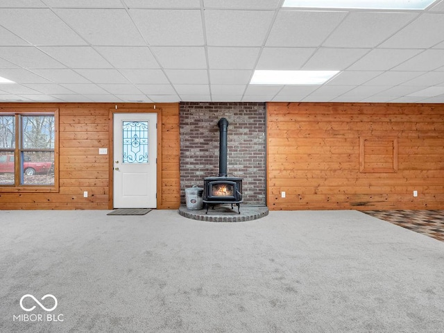 unfurnished living room featuring a paneled ceiling, wooden walls, carpet floors, and a wood stove