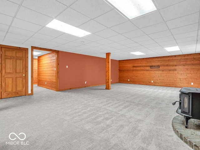 basement with a paneled ceiling, a wood stove, light carpet, and wooden walls