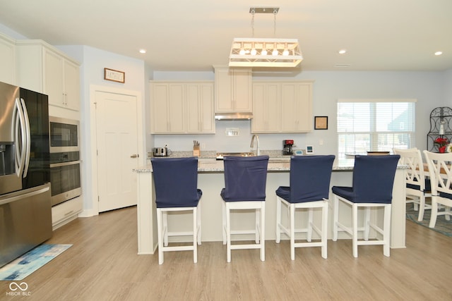 kitchen with light stone countertops, appliances with stainless steel finishes, a center island with sink, light hardwood / wood-style flooring, and hanging light fixtures