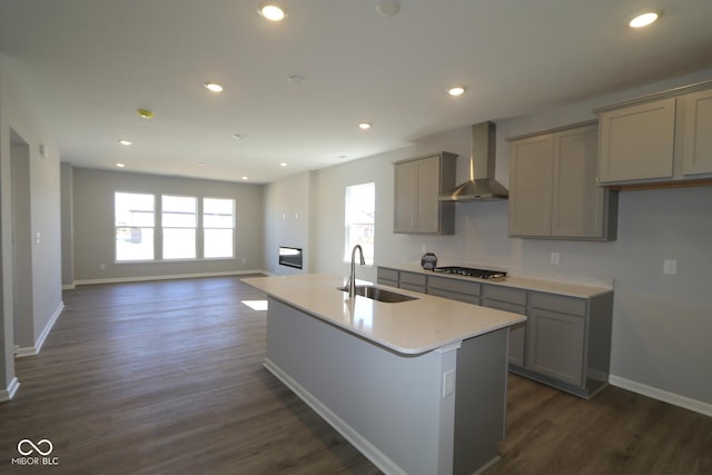 kitchen with a center island with sink, a sink, gray cabinetry, stainless steel gas stovetop, and wall chimney exhaust hood