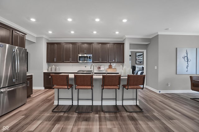 kitchen with a kitchen island with sink, dark brown cabinetry, wood finished floors, appliances with stainless steel finishes, and backsplash