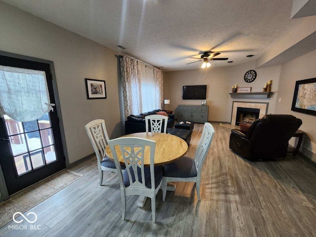 dining area with a tile fireplace, wood-type flooring, a textured ceiling, and ceiling fan