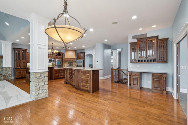 kitchen featuring premium range hood, stainless steel microwave, a notable chandelier, pendant lighting, and a kitchen island with sink