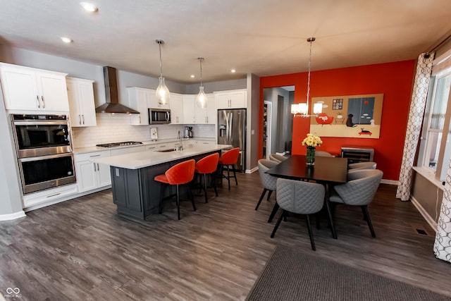 kitchen with appliances with stainless steel finishes, pendant lighting, white cabinetry, a kitchen island with sink, and wall chimney range hood