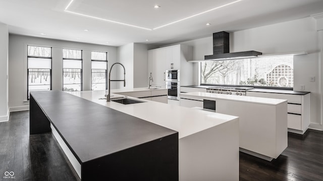 kitchen with a large island with sink, wall chimney exhaust hood, white cabinetry, stainless steel appliances, and sink