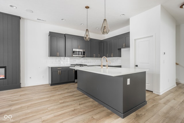 kitchen with hanging light fixtures, stainless steel appliances, a center island with sink, light hardwood / wood-style floors, and decorative backsplash