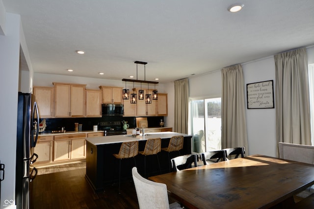 kitchen with hanging light fixtures, electric range, a kitchen island with sink, stainless steel refrigerator, and light brown cabinets