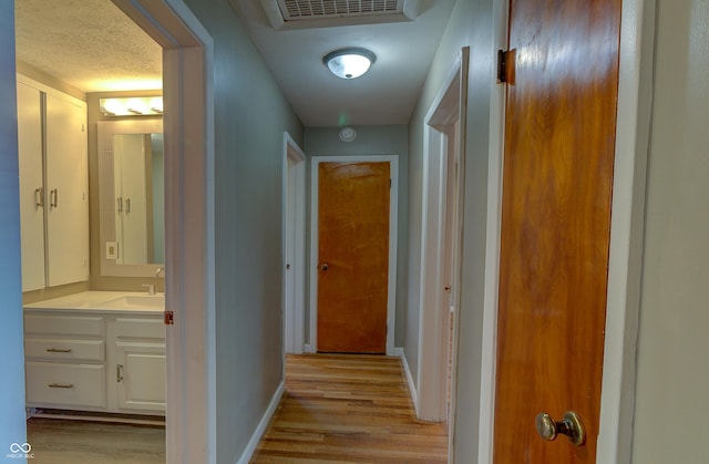 hall with visible vents, light wood-style floors, a sink, a textured ceiling, and baseboards