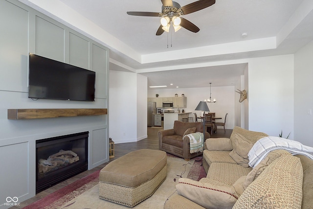 living room featuring a raised ceiling, ceiling fan with notable chandelier, wood finished floors, a glass covered fireplace, and baseboards