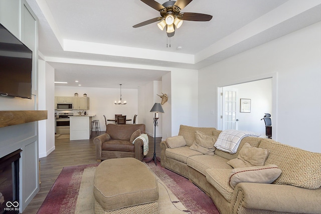 living room featuring a raised ceiling, dark wood-style floors, a fireplace, and ceiling fan with notable chandelier