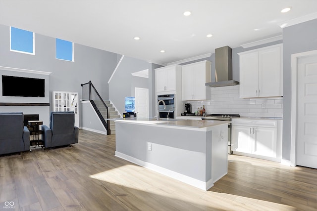 kitchen featuring appliances with stainless steel finishes, wall chimney exhaust hood, light hardwood / wood-style flooring, white cabinetry, and a center island with sink