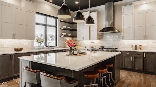 kitchen featuring wall chimney exhaust hood, a center island with sink, hanging light fixtures, and light stone countertops