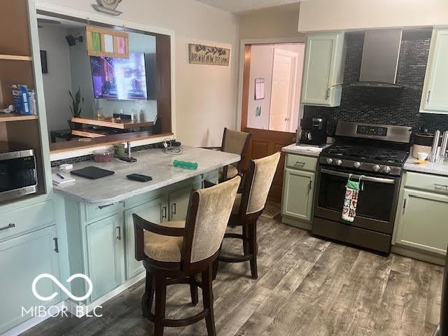 kitchen with wall chimney range hood, decorative backsplash, wood-type flooring, appliances with stainless steel finishes, and green cabinetry