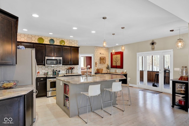 kitchen featuring an island with sink, appliances with stainless steel finishes, decorative light fixtures, dark brown cabinets, and light stone counters