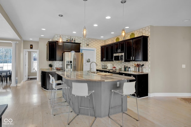 kitchen featuring pendant lighting, appliances with stainless steel finishes, an island with sink, decorative backsplash, and decorative columns